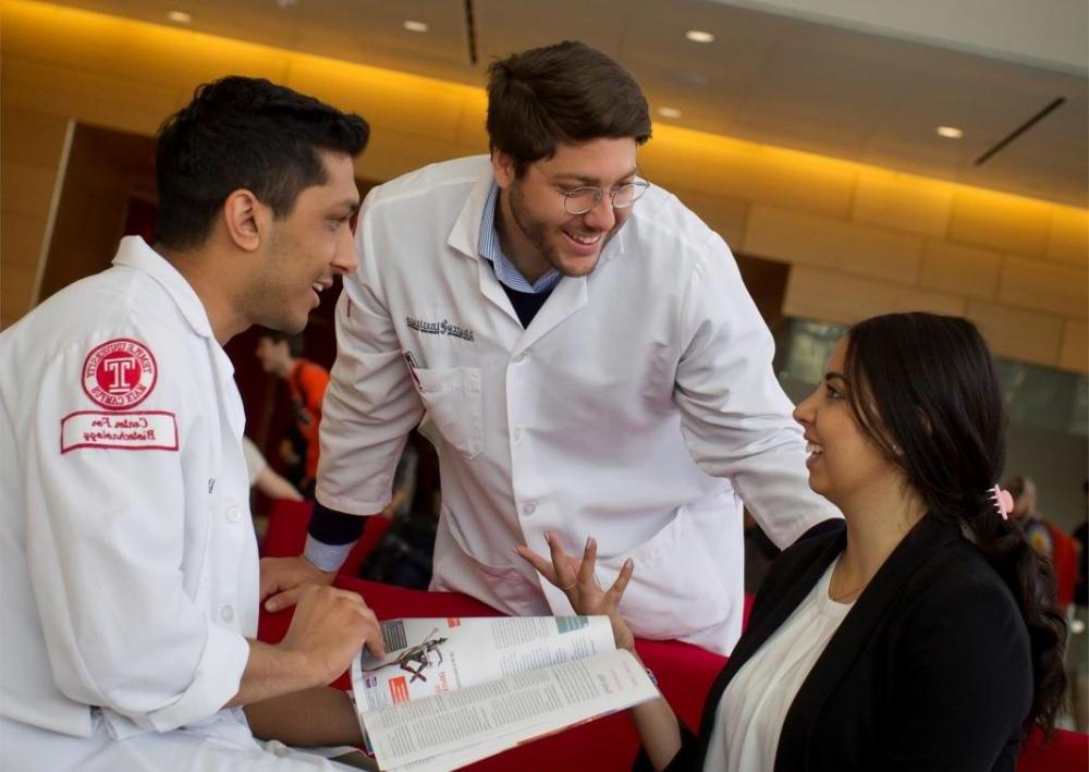 Two students in white lab coats talk with another student.